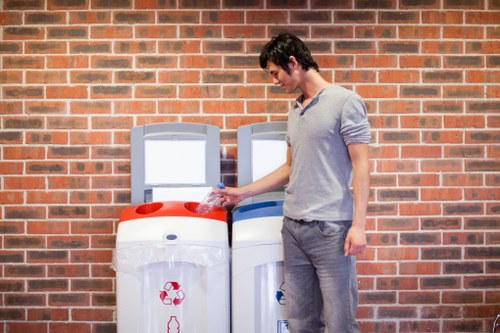 A young man with dark hair, wearing a light grey long-sleeved top and grey trousers, is standing outdoors in front of a red brick wall. He is holding a plastic water bottle in his right hand, which he appears to be discarding into a white recycling bin with a red recycling symbol. Next to him is a similar bin with a blue symbol, likely for general waste. Both bins have simple, rounded tops with coloured sections to denote recycling and waste separation, and are positioned on a paved surface. The environment suggests a residential or communal area, possibly in the Blackheath area, established with a straightforward setup for waste disposal. The image is well-lit, indicating daylight, and the overall scene reflects responsible rubbish separation activities consistent with local rubbish removal services offered by Blackheath House Clearance.